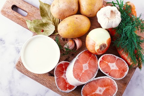 Potatoes, salmon, garlic, onion, and herbs are placed on a wooden cutting board. These ingredients