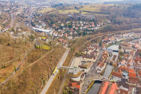 Panoramic picture of a city with river and railroad tracks, surrounded by wooded hills, acceptance