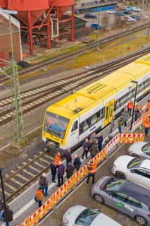 A yellow train at a train station with people and parked cars on the side, acceptance of the
