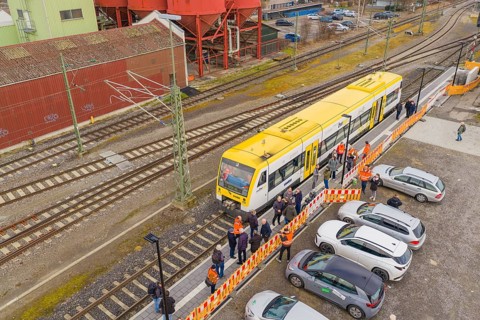 Yellow train at a train station with parked cars and people, red industrial structure, acceptance
