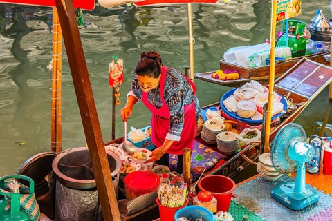 Elderly woman wearing mask offers food on her wooden boat, Damneon Saduak floating markets channel,