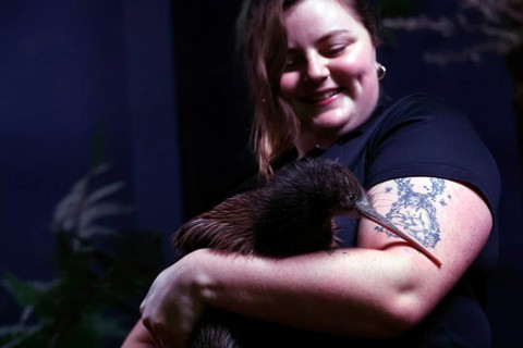 Woman smiling as she gently hugs a kiwi, visible a tattoo on her arm, Franz Josef Glacier, West