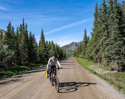 Young woman riding a bicycle on a dirt road through taiga, mountainous landscape, Denali Park Road,