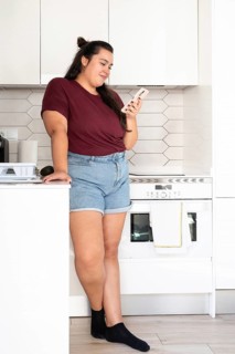 A curvy woman in a casual burgundy top and denim shorts is leaning against a kitchen counter,