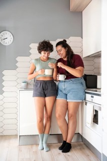 A happy lesbian couple stands in a kitchen, chatting and sharing a morning coffee. They enjoy