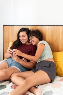 Two lesbian women sitting on a bed, enjoying a relaxed moment with a smartphone and a notebook.