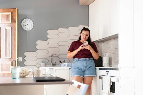 A curvy woman in a casual outfit stands in a modern kitchen, intently using her smartphone. The