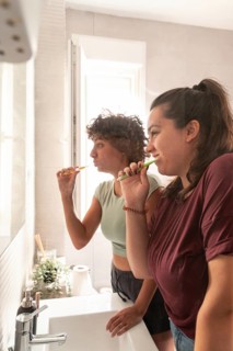 A lesbian couple shares a morning routine in the bathroom, with one applying makeup and the other