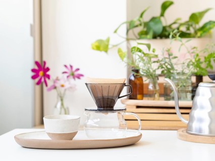 Modern pour over coffee brewing setup sits on a white table, with houseplants and flowers adding a