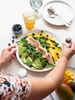 Woman seasoning a colorful healthy salad with chicken, mango, avocado, blueberries and lamb's