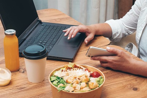 A woman is working on a laptop at home, there is food on the table, salad, coffee and juice,