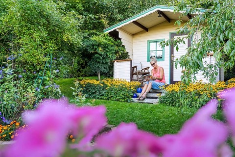 A woman sits relaxed on the small veranda of a garden shed and reads. Blooming summer flowers in