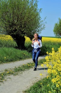 Young woman with backpack walking on a countryroad with willow trees and field of rape in Skivarp,