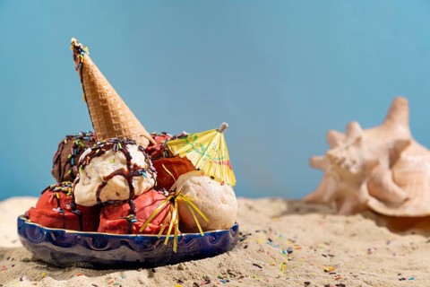 Various flavours of ice cream with chocolate sauce, crumble and parasol on sand with shells