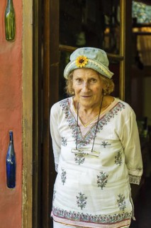 Elderly woman wearing a denim hat with a sunflower decoration and a white tunic smiles warmly at