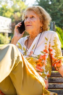 Senior woman sitting comfortably on a park bench, making a phone call and enjoying the warm summer