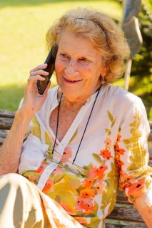 Elderly woman with glasses hanging from a string around her neck is sitting on a wooden bench