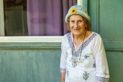 Elderly woman smiling while leaning against a light green wooden door, wearing a light blue hat
