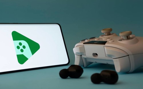 A smartphone with a Google Play Games app icon next to a controller and earbuds on a blue surface