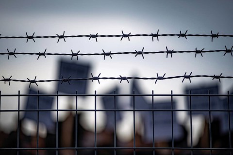 Barbed wire fence with blurred protesters and signs in background during border or prison