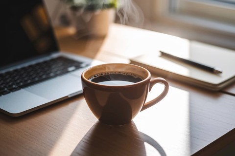 Cup of steaming coffee on desk. Cozy workspace with notebook in background, Generative ai, AI