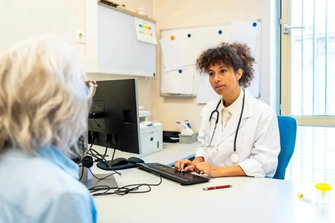 Female doctor using computer and listening to senior patient describing symptoms in hospital room