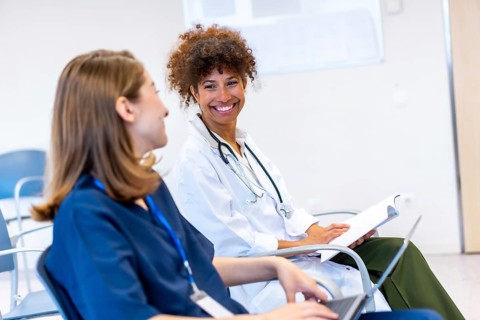 Two female doctors are sitting and smiling while discussing medical information during a conference