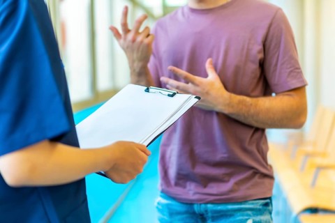 Young man explaining his symptoms using gestures to a nurse holding a medical chart in a hospital