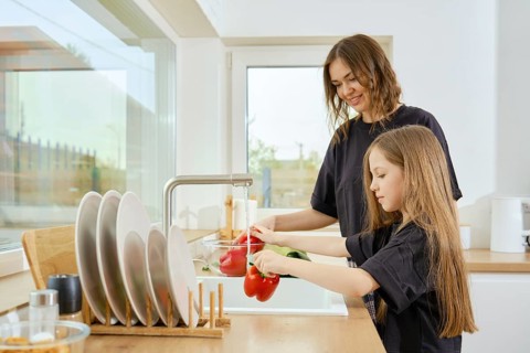 A mother watches her daughter wash vegetables under running water in the kitchen