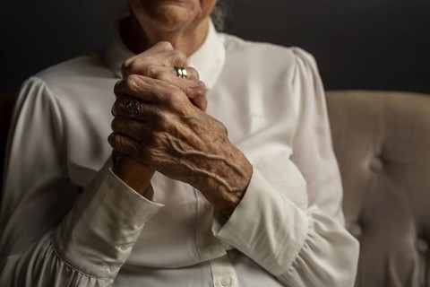 90 year old woman in white blouse holding folded hands, conveying thoughtfulness in a dim light,