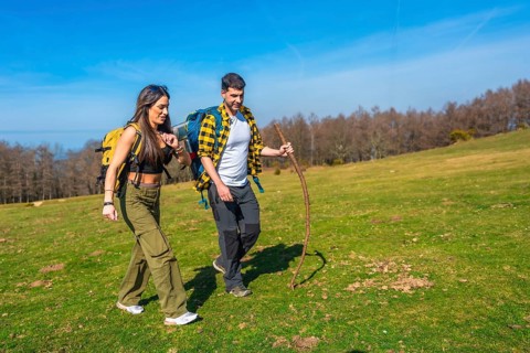 Young couple walking hand in hand across a vibrant green meadow, carrying backpacks and using a