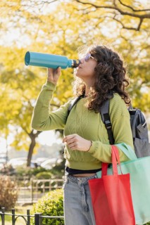Woman with curly hair and sunglasses drinks from a blue water bottle in a park. She carries