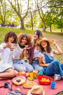 Four cheerful young women enjoying a sunny picnic in the park, happily taking a selfie with their