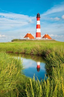 The Westerheversand lighthouse under a blue sky with white clouds reflected in a tideway,