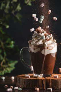 Marshmallows and cocoa powder fall onto a cup of hot chocolate and whipped cream on a wooden board