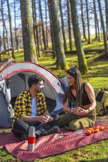 Couple playing cards near tent and backpack in forest during camping trip, enjoying sunny day in