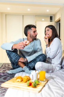 Young couple wearing pajamas sitting in bed eating donuts for breakfast, with croissants, orange
