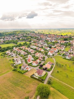 Residential area with densely built houses and streets, surrounded by fields under a slightly