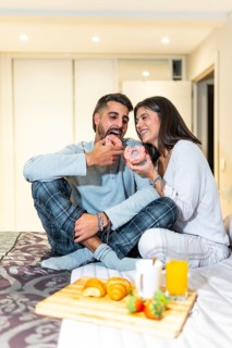 Young couple wearing pajamas, sitting in bed and eating breakfast, including donuts, croissants,