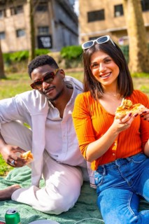 Happy interracial couple eating pizza outdoors, enjoying a relaxing picnic in a sunny urban park