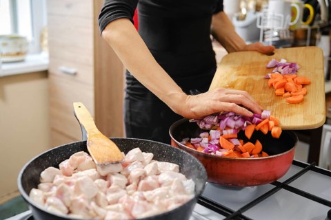 Unrecognizable woman cooking chicken pieces in one pan while pouring chopped onions and carrots to