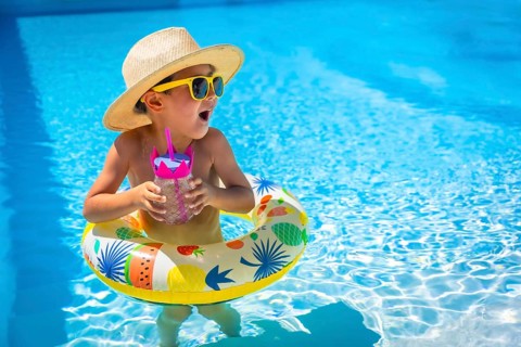 Playful caucasian male kid in sun hat and sunglasses on a pool with a glass of water standing
