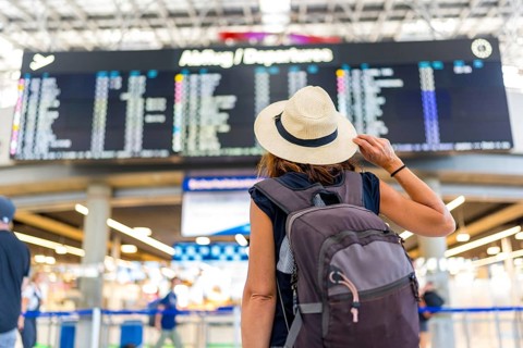 A woman wearing a straw hat and a backpack is looking at a large airport screen. She is waiting for