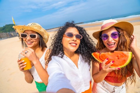 Three women are smiling and holding watermelons on a beach. They are wearing straw hats and