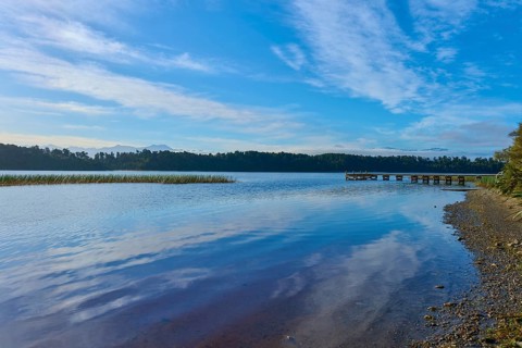 A jetty and shingle beach stretch along the shore of a tranquil lake under blue skies, Lake