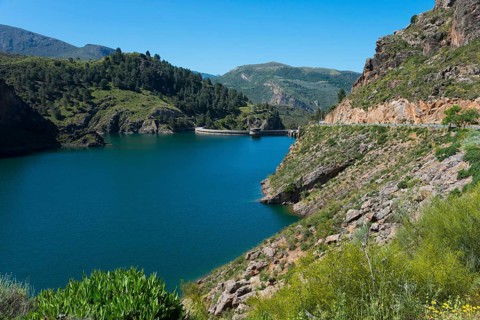 A tranquil lake in a mountainous landscape with green hills and rocks, under a clear blue sky,