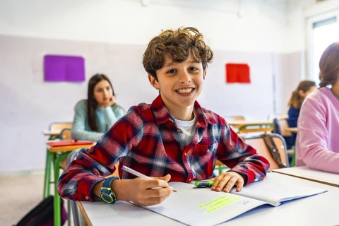 Happy elementary school student smiling and writing in notebook during class, with classmates in