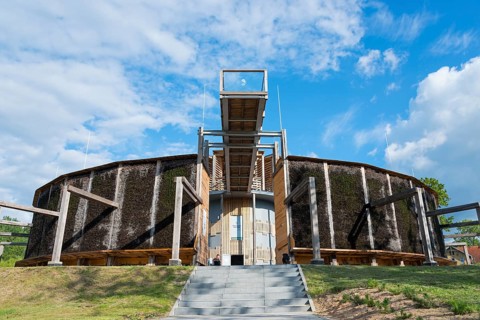 Round building made of wood and metal with stairs, blue sky and green landscape in the background,