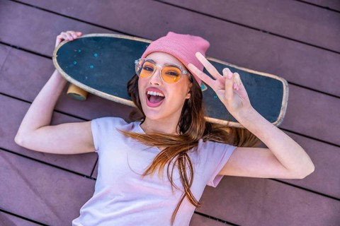 Happy young woman lying on a wooden floor with her skateboard showing peace sign, wearing pink