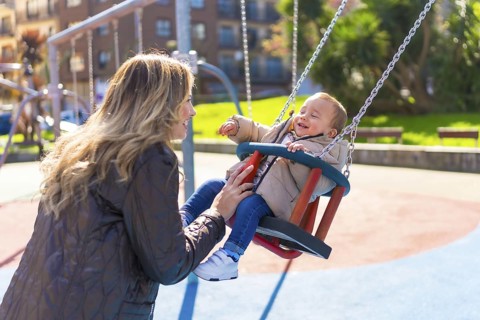 Happy mother pushing her laughing baby son on a swing, enjoying a sunny autumn day at the